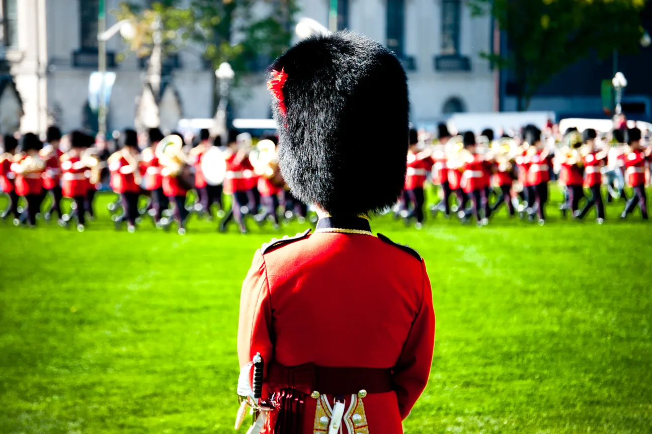 Ceremonial Guard soldier in red tunic and bearskin cap standing before the marching band on Parliament Hill's lawn